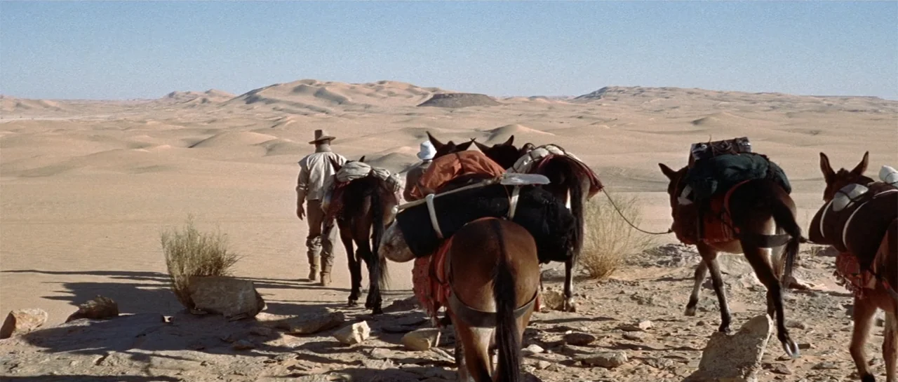 Legend of the Lost (1957) movie still: wide — A vast sandy expanse under a clear sky with rolling dunes in the distance. Two men lead a small train…
