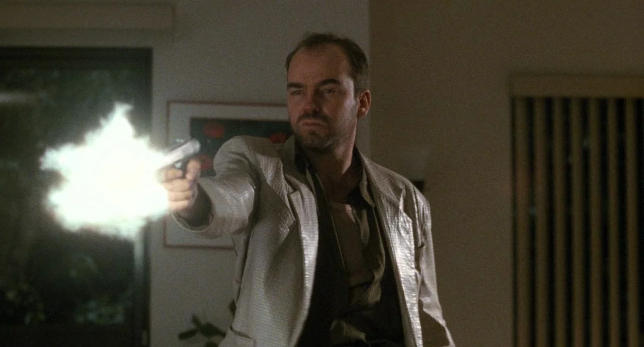 One False Move (1991) movie still: medium — A man in a cream jacket stands inside a dimly lit domestic space. He extends his arms to fire a handgun…