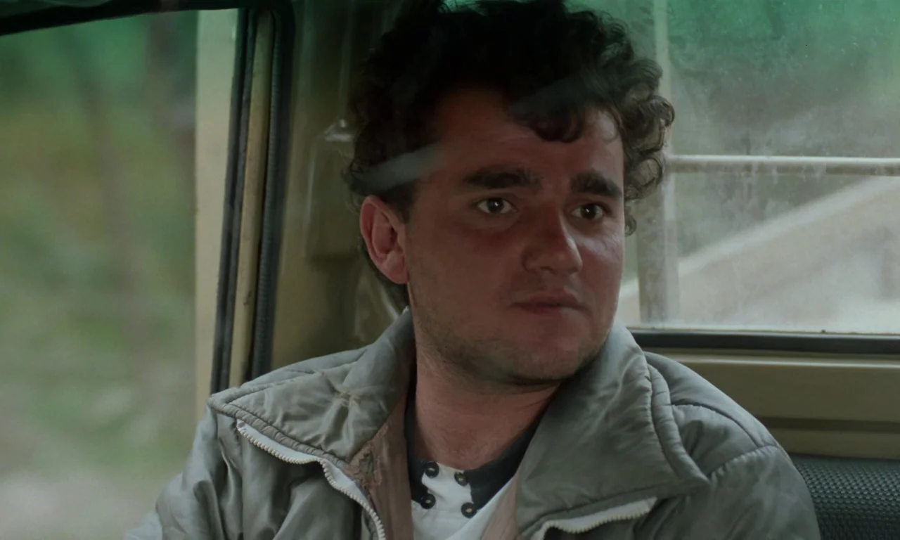 Through The Olive Trees (1994) movie still: medium — The man sits inside a vehicle with tan paneling and window frames. The man looks ahead while…