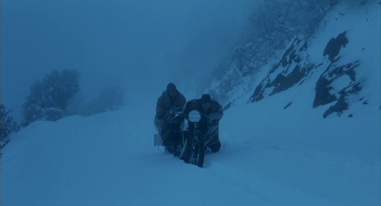 The Motorcycle Diaries (2004) movie still: wide — A snowy, desolate mountain pass with rocky slopes and thick snow coverage. Two men push a…