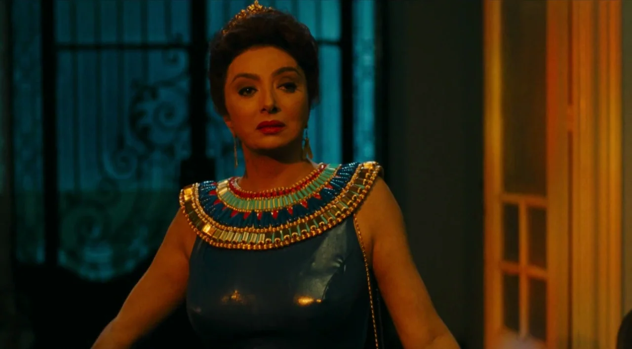The Other Conquest (1999) movie still: medium — A woman stands in a dimly lit interior space with a door behind her. She wears an elaborate blue…
