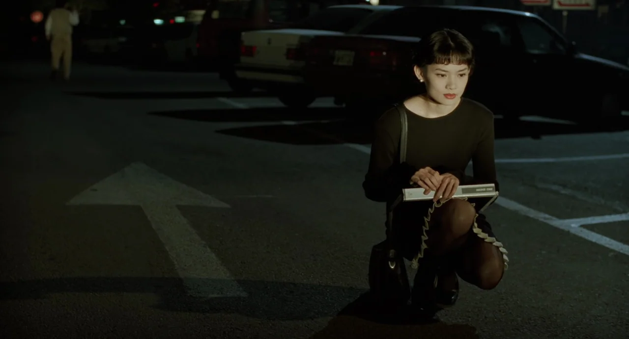 A Confucian Confusion (1994) movie still: medium — An asphalt parking lot at night features a white painted arrow on the ground. A young woman in a…