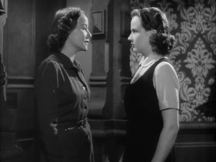 The Seventh Victim (1943) movie still: medium — Two women stand in an indoor corridor with patterned wallpaper and dark wood panels. They face each…