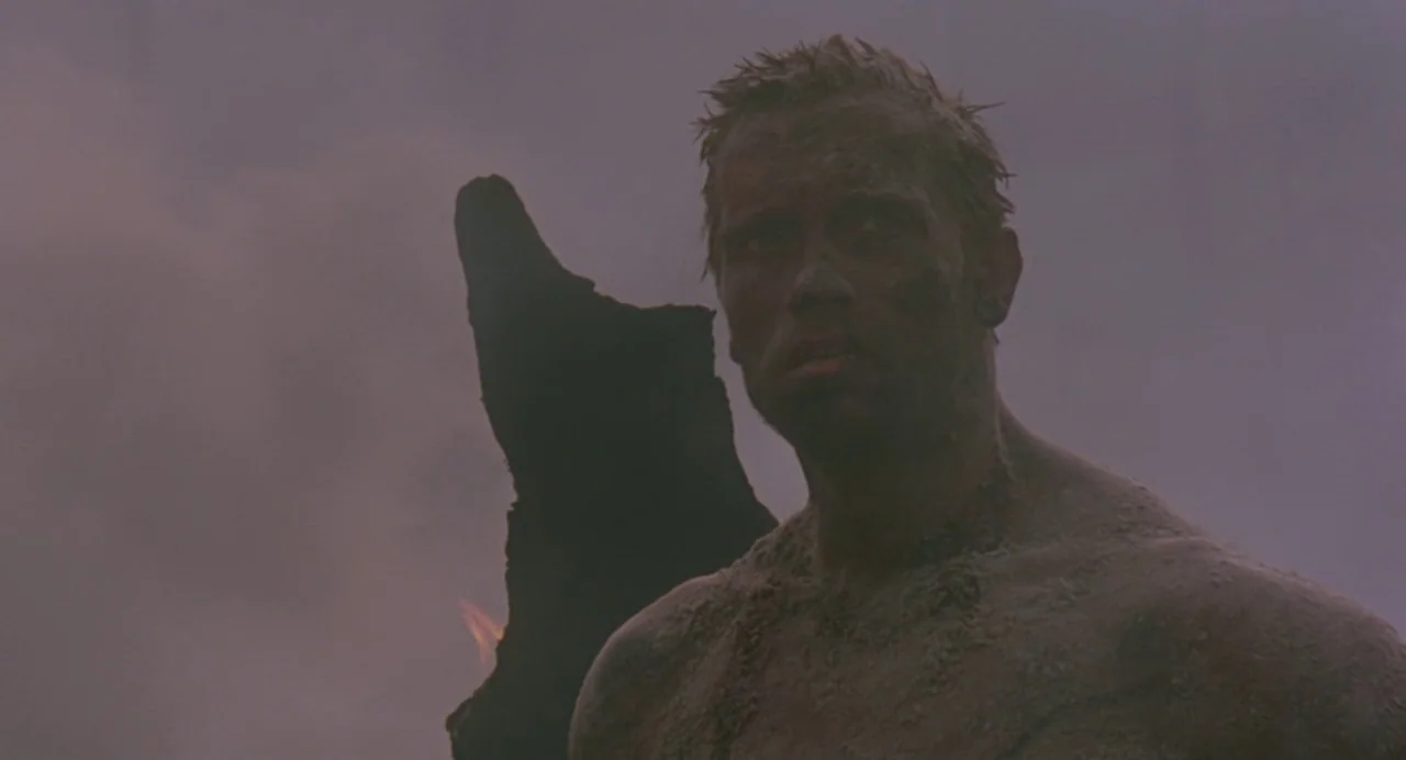 Predator (1987) movie still: medium — A dark, hazy beach environment under a dim, overcast night sky. A muscular man covered in debris stands in the…