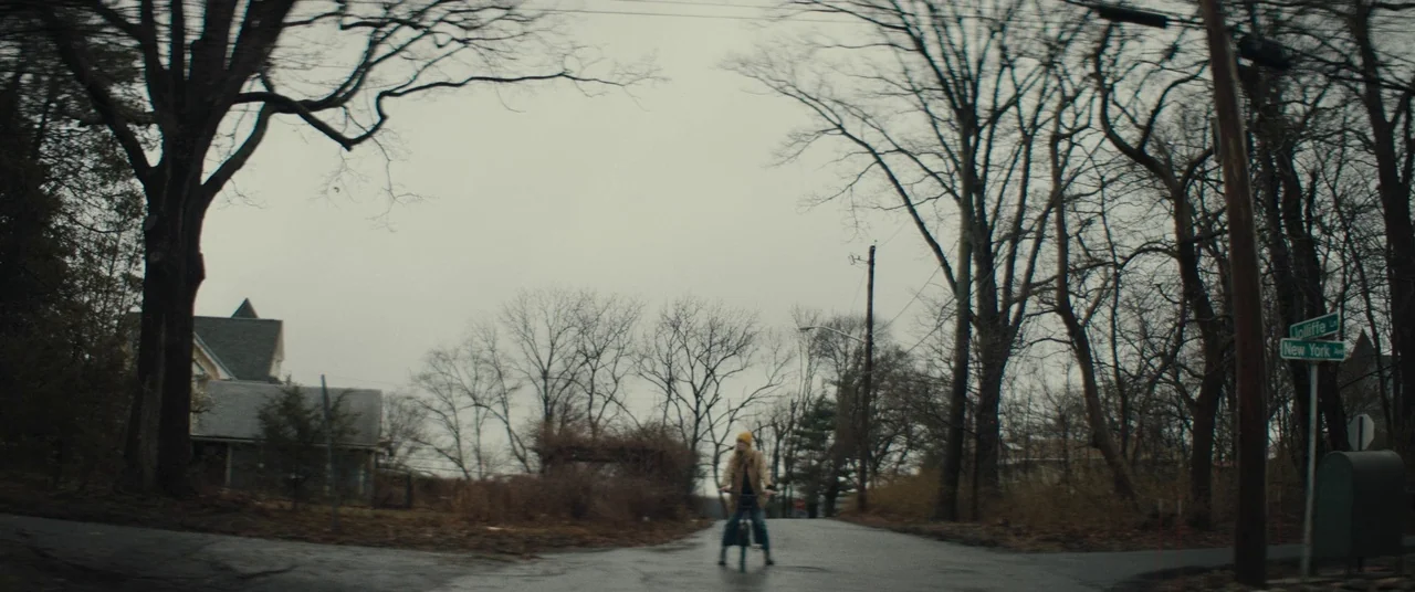 I Think We’re Alone Now (2018) movie still: wide — A suburban intersection features bare trees under a heavy, overcast winter sky. A lone person on…