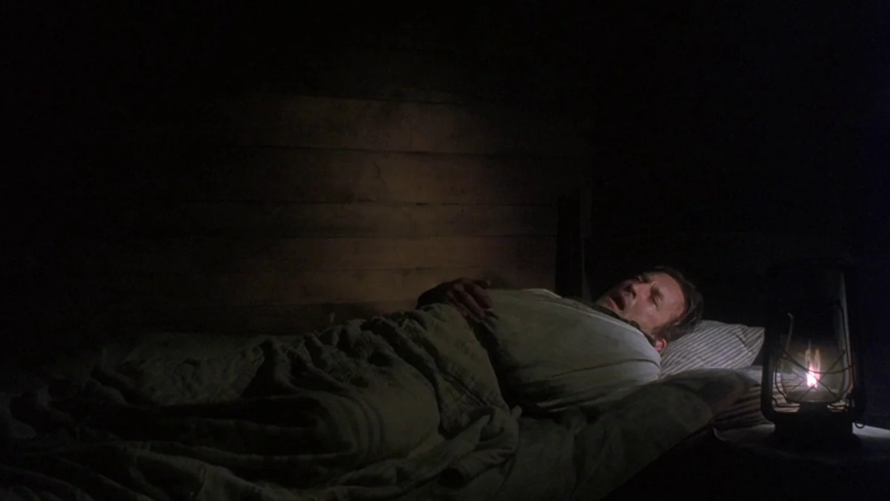 Matewan (1987) movie still: medium — A man lies in a bed against a wooden-paneled wall inside a dark room. He is sleeping while an oil lamp flickers…