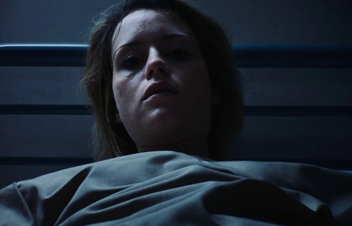 Unsane (2018) movie still: closeup — A dimly lit hospital room features a metallic headboard frame in the background. A woman lies flat on a bed,…