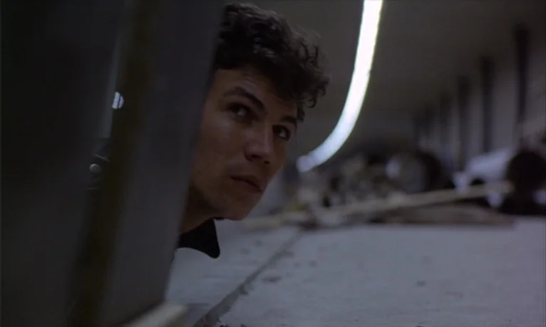Spetters (1980) movie still: closeup — An interior space contains a concrete surface and a vertical structure in the foreground. A young man with…
