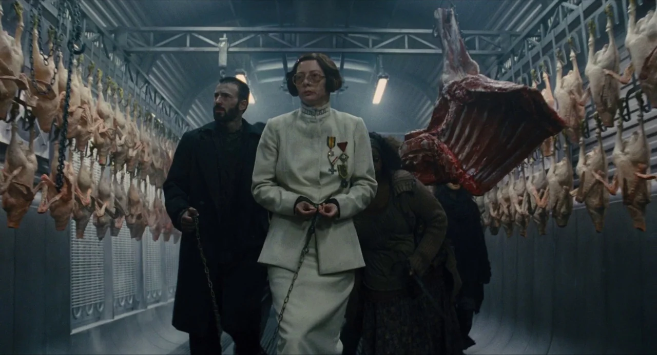 Snowpiercer (2013) movie still: fullbody — A cramped industrial train car interior features rows of hanging raw chicken carcasses. Several people…