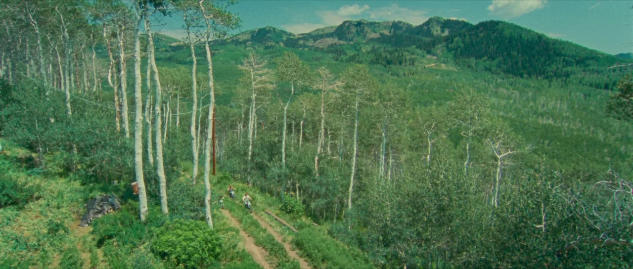 Riddle of Fire (2023) movie still: wide — A dense forest of white-barked trees covers a steep mountainside under a bright sky. Three cyclists ride…