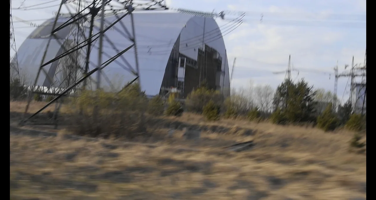 Chernobyl's Café (2016) movie still: wide — A large metallic dome structure stands behind tall electricity pylons in a dry field. Sparse trees and…