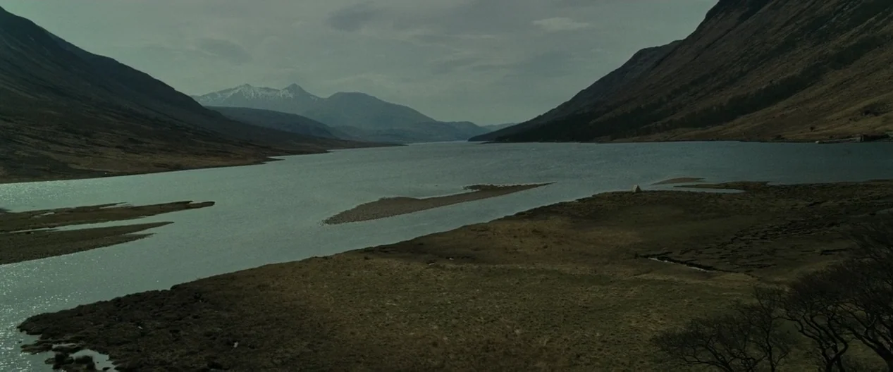 A vast loch sits between tall grass-covered mountains under a heavy overcast sky. Small gravel islands emerge from the water with snow visible on the distant peaks.