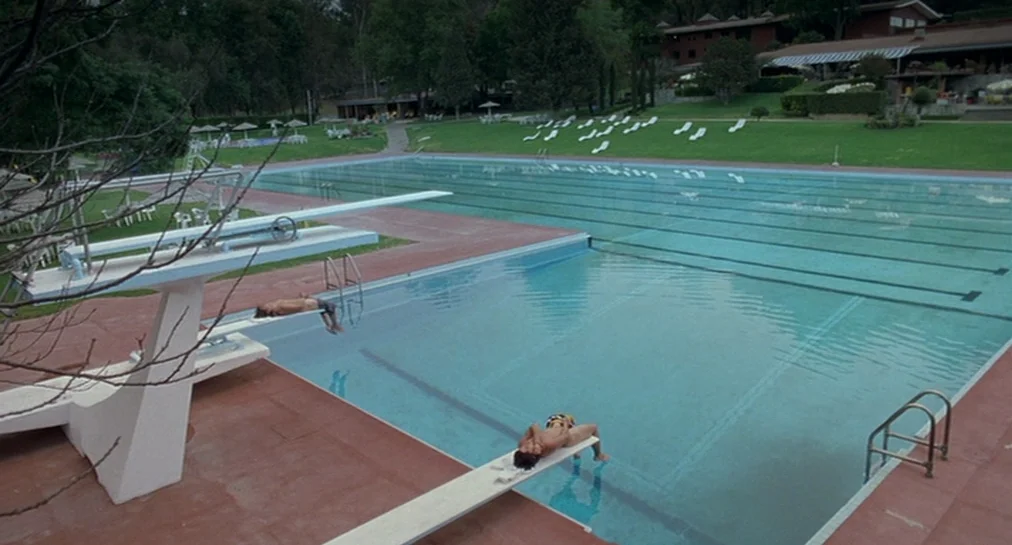 Y Tu Mamá También (2001) movie still: wide — An empty public swimming pool features tiered diving boards in a park-like setting. Two individuals lie…