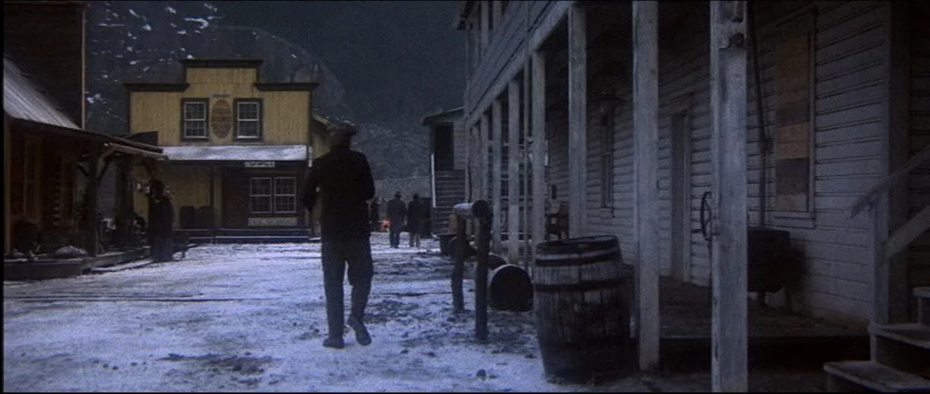 McCabe & Mrs.Miller (1971) movie still: wide — A snow-covered street in a wooden western town features buildings with porches at night. A man walks…