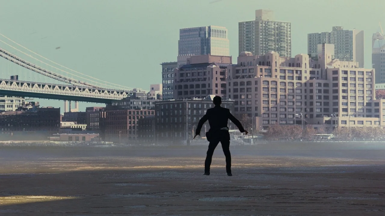 The Dark Knight Rises (2012) movie still: wide — A man stands on a weathered concrete pier facing a large suspension bridge. Dense city architecture…