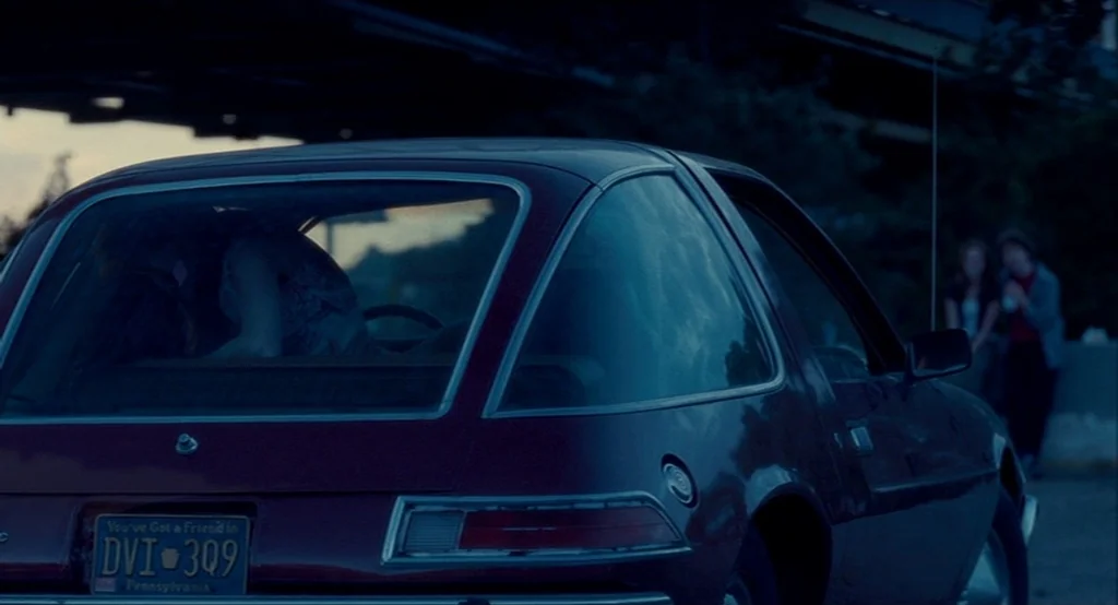 Adventureland (2009) movie still: medium — A dark 1970s AMC Pacer sits under a concrete overpass during dusk. A person sits in the driver seat while…
