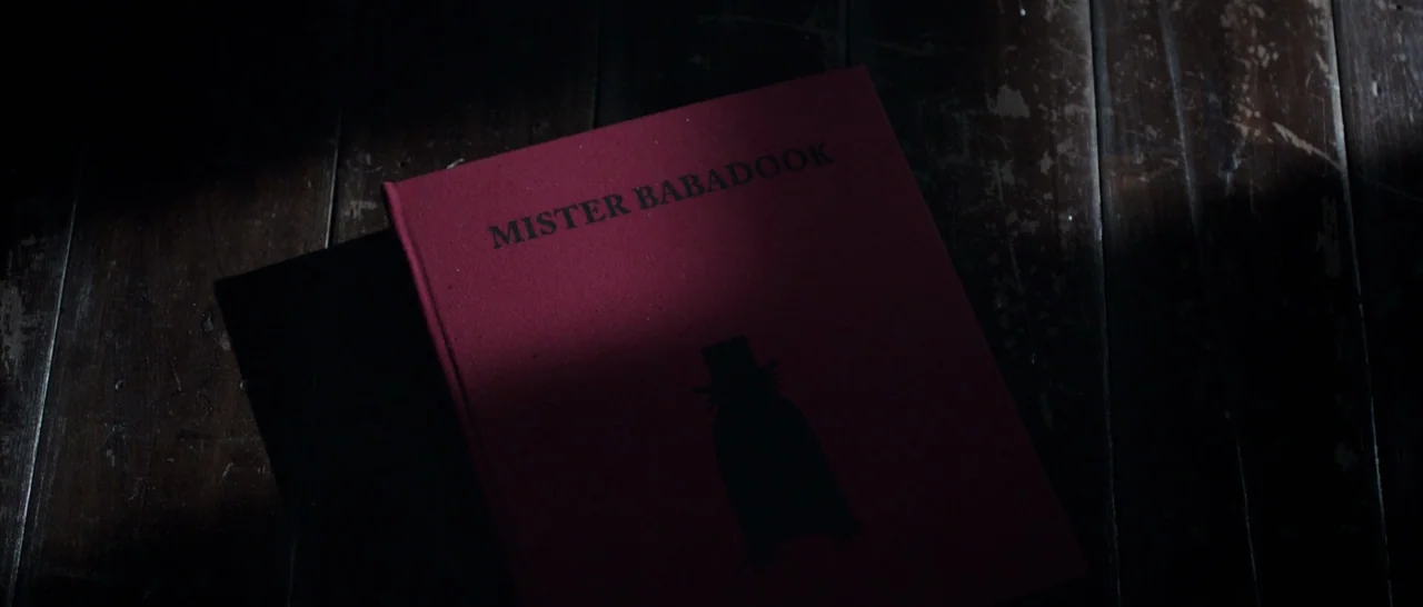 The Babadook (2014) movie still: closeup — A red book with black text and a silhouette graphic rests on dark wood. A sharp shaft of light…