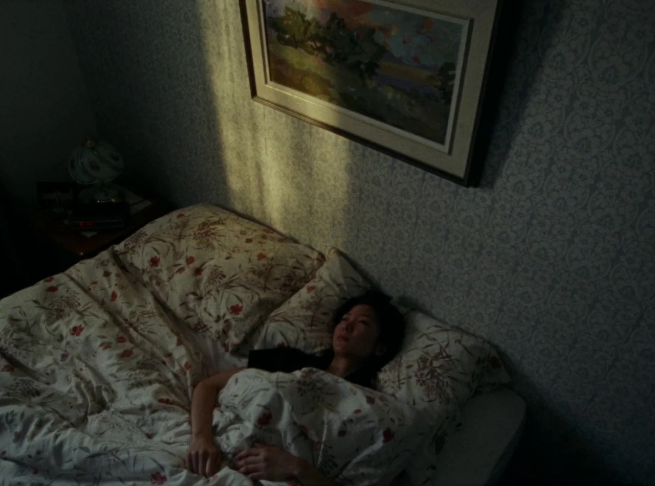 Riceboy Sleeps (2022) movie still: medium — A dark bedroom with patterned wallpaper is illuminated by a shaft of morning light. A woman lies in bed,…