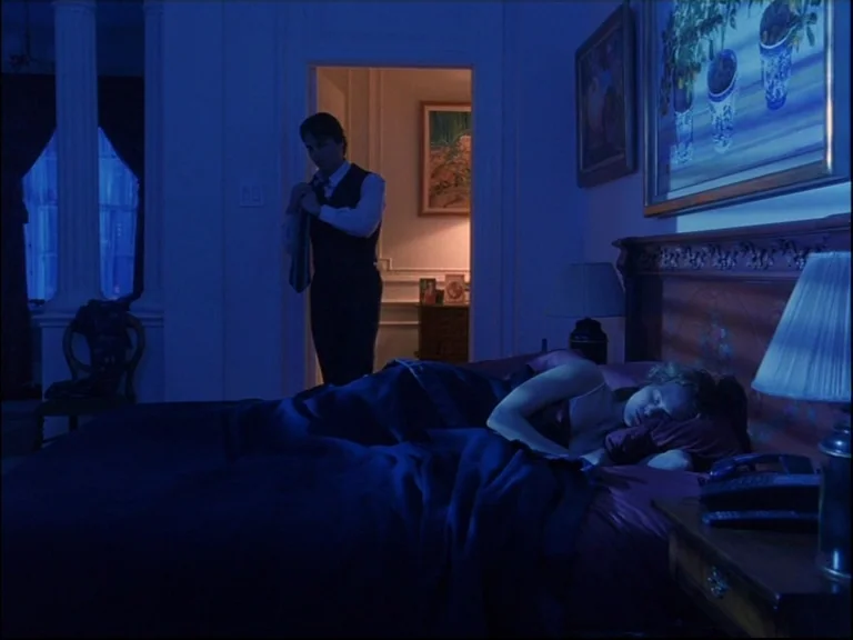 Eyes Wide Shut (1999) movie still: wide — A dark, blue-lit bedroom contains a bed with a woman asleep in it. A man stands near an open doorway…