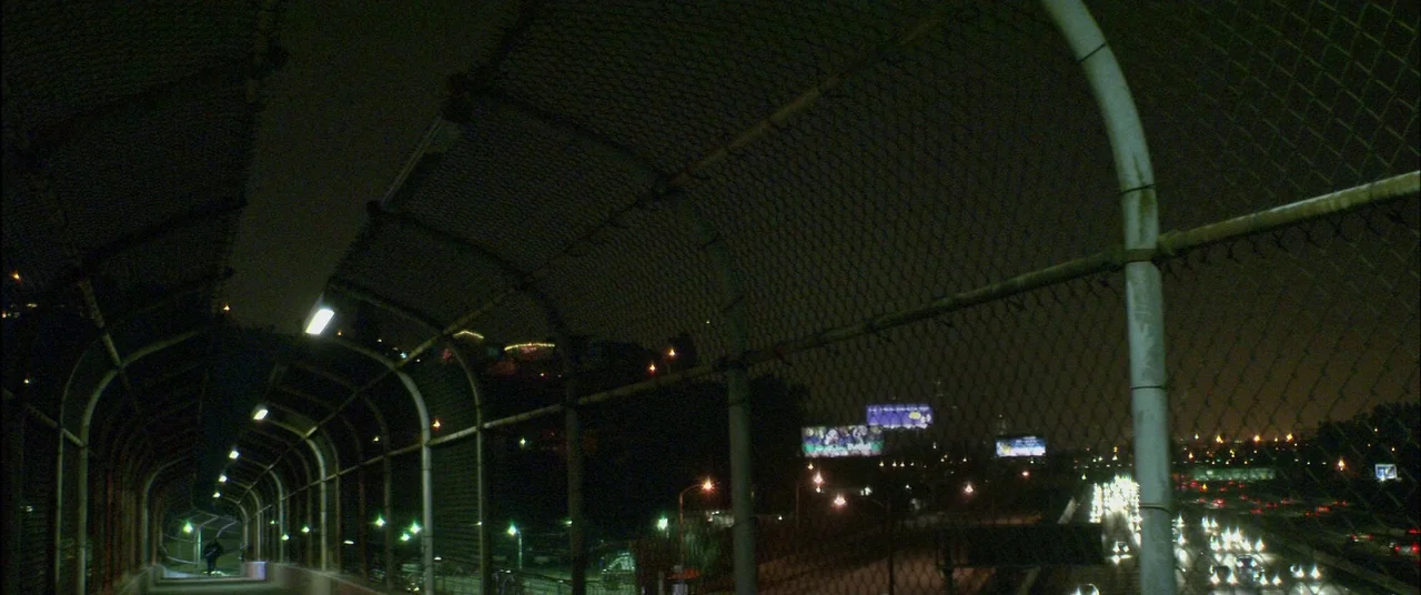 Collateral (2004) movie still: establishing — A fenced-in pedestrian overpass spans across a dark multi-lane highway at night. Small bright lights…