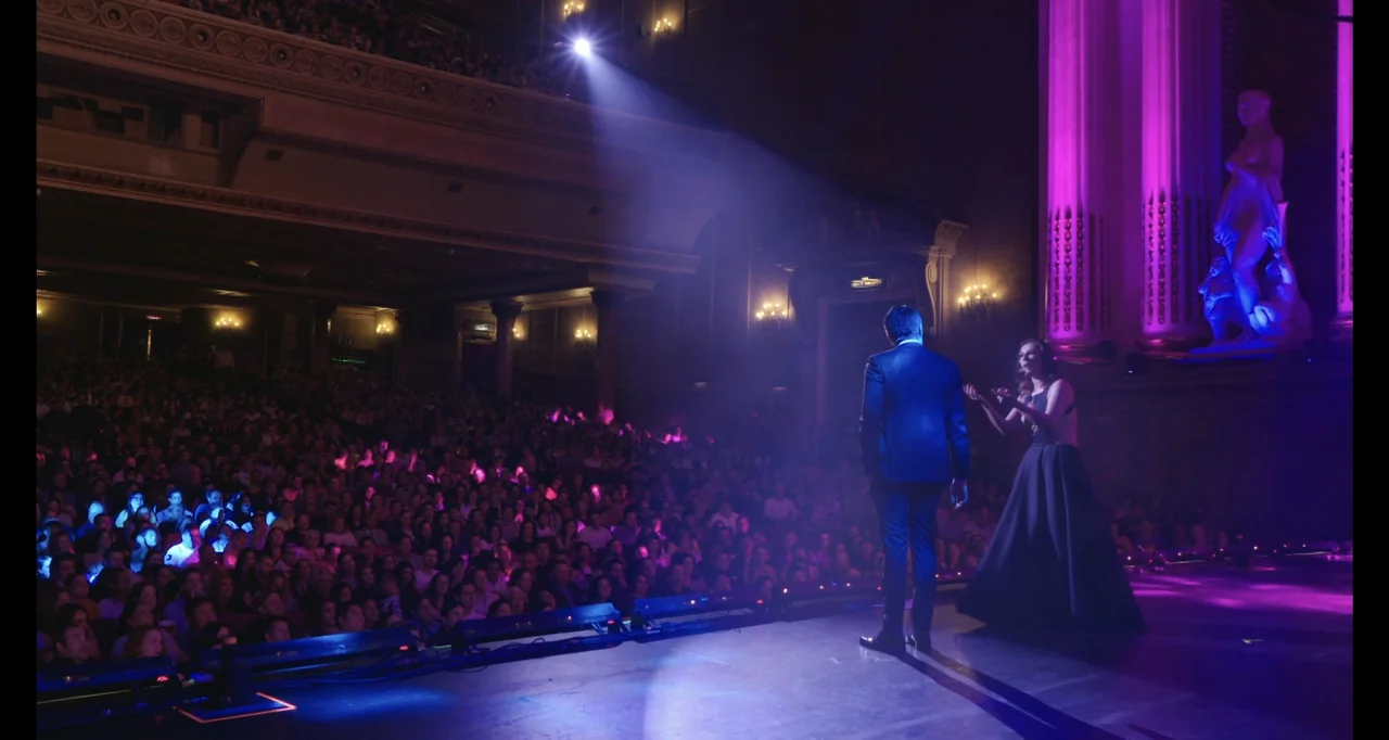 Paired Up (2019) movie still: wide — A large theater auditorium is filled with an audience watching a performance on stage. A man and a woman in…