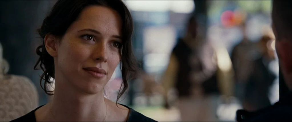 The Town (2010) movie still: closeup — An outdoor public area with blurred figures in the background. A woman with dark hair looks towards an unseen…