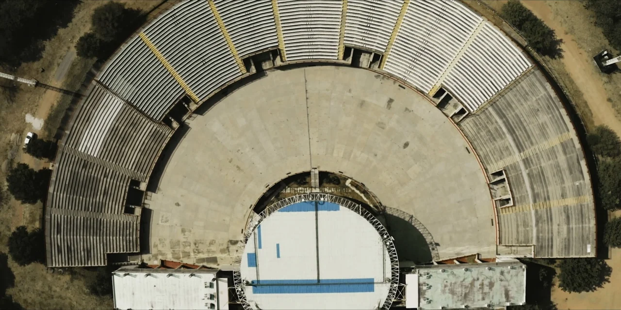 Leyenda Viva (2022) movie still: establishing — An empty semicircular concrete amphitheater features tiered seating rows arranged around a circular…