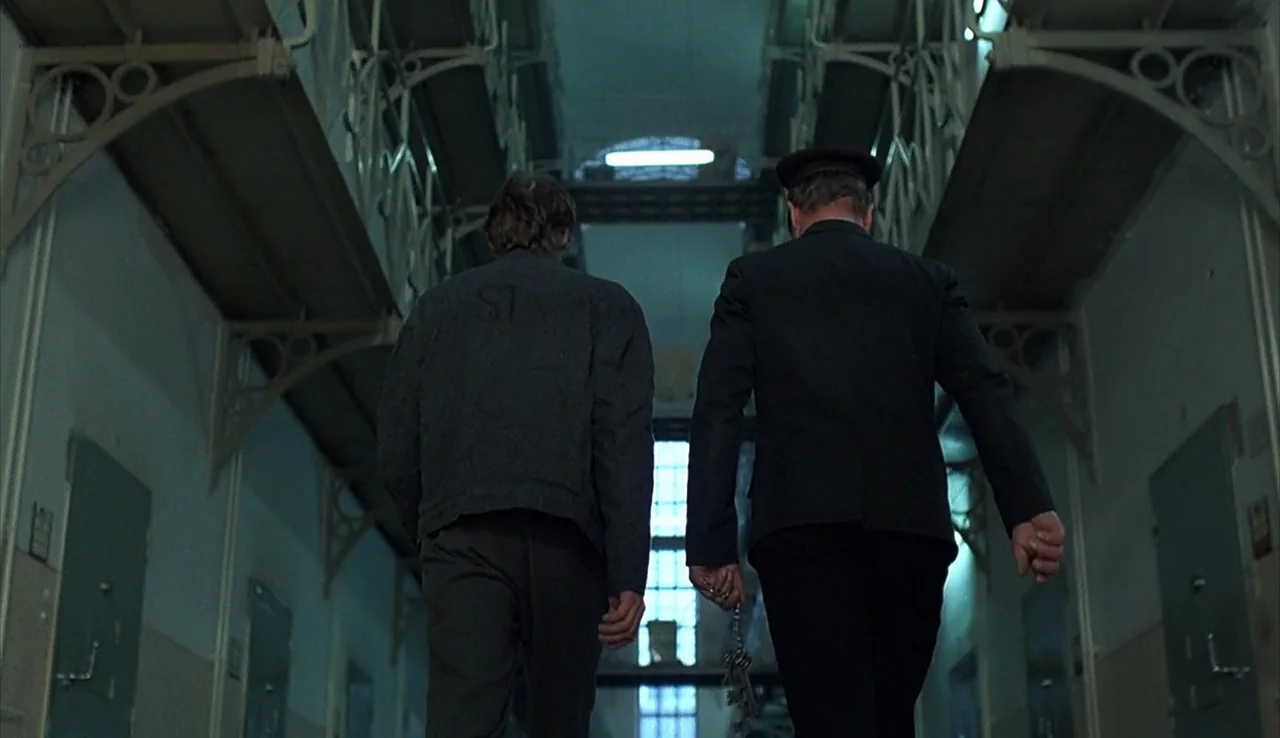 Angst (1983) movie still: fullbody — A long, industrial prison corridor with rows of cell doors and iron catwalks above. A man in a prisoner jacket…