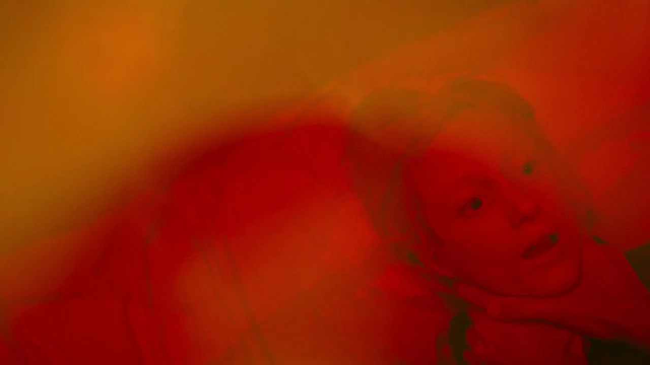 Possessor (2020) movie still: closeup — A dimly lit room bathed in an intense monochromatic red light. A person lies down in the foreground with…
