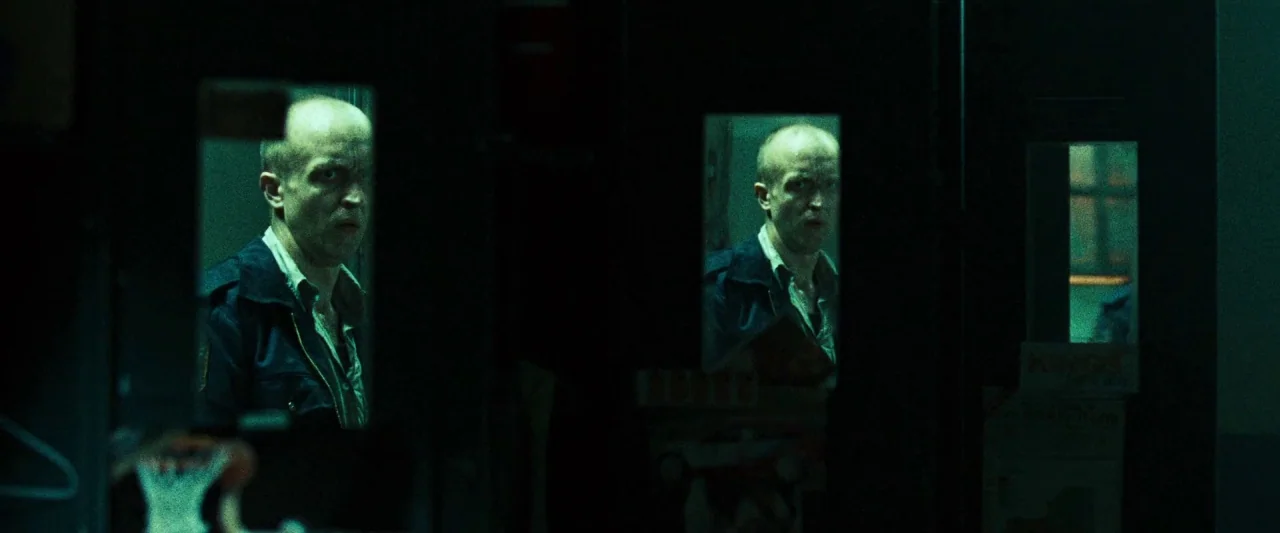 Mirrors (2008) movie still: medium — A dark hallway features heavy doors with inset glass panels reflecting a man's face. The man peers through the…