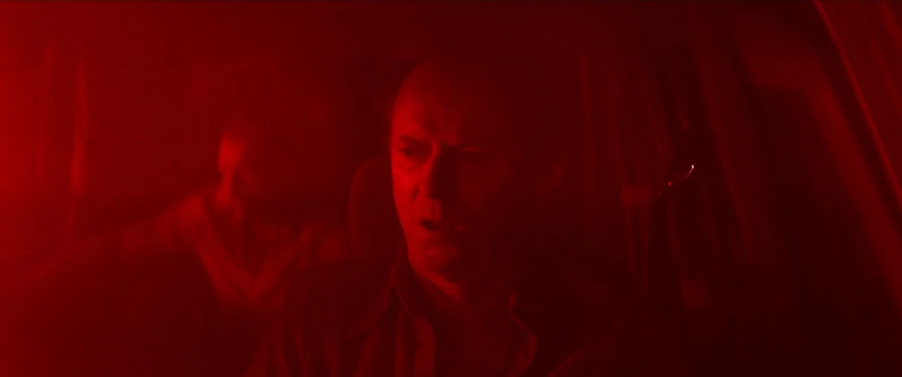 Mandy (2018) movie still: medium — A dark car cabin is bathed in intense, immersive monochromatic red light. A middle-aged man sits behind the wheel…