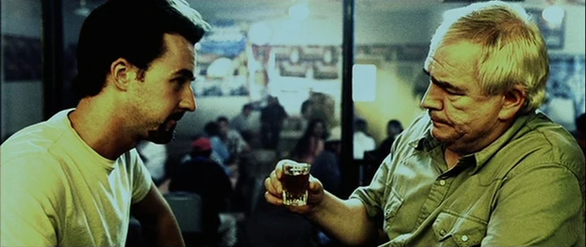 25th Hour (2002) movie still: medium — A dimly lit bar with blurry patrons sitting in the background darkness. An older man offers a small shot…