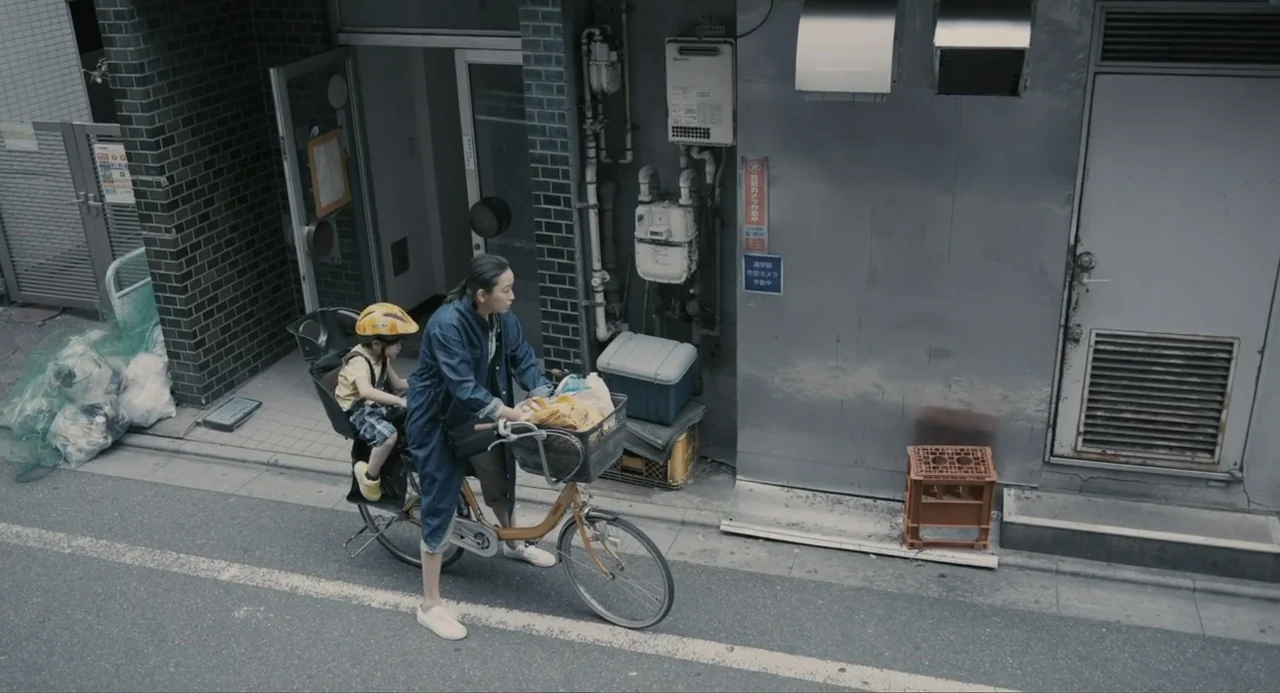 Elles (2018) movie still: medium — A narrow Japanese city street with a grey metal wall and dark brick building. A woman in a denim coat rides a…