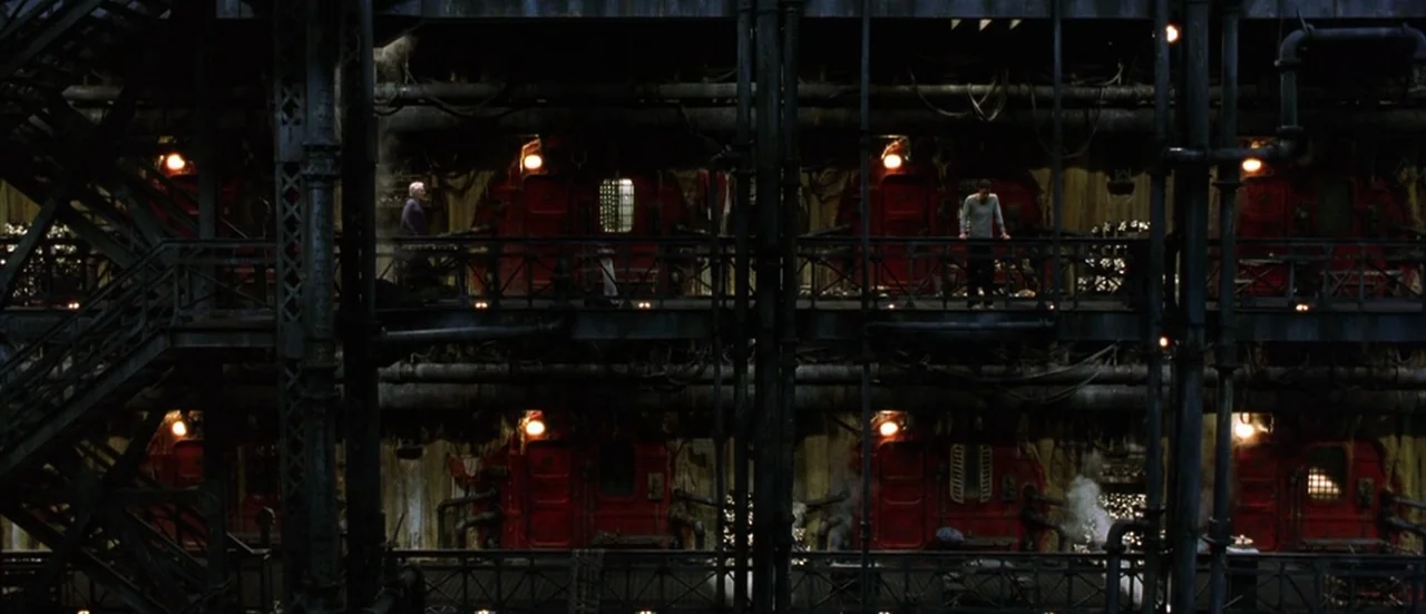 The Matrix Reloaded (2003) movie still: wide — A multi-level industrial steel catwalk structure features red metal door segments and exposed pipes.…