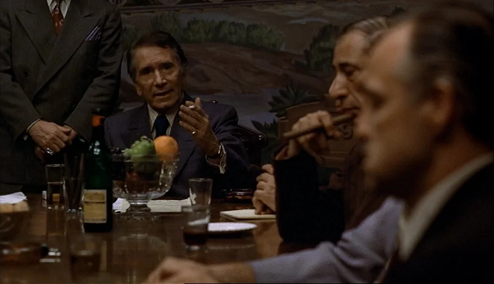 The Godfather (1972) movie still: medium — A wood-paneled room contains a long table with glasses, bottles, and fruit bowls. Three men in suits sit…