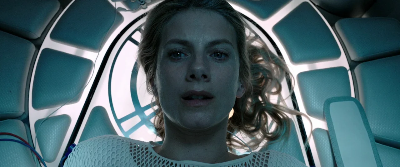 Oxygen (2021) movie still: closeup — The interior of a futuristic, white-padded medical chamber with glowing circular accent lighting. A woman lies…