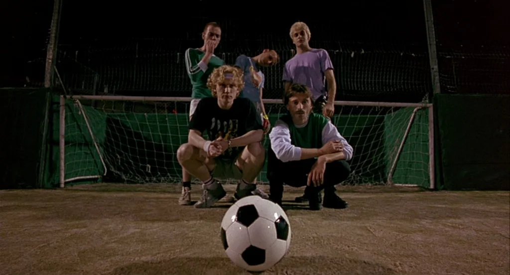 Trainspotting (1996) movie still: fullbody — A fenced-in soccer field at night with concrete ground and dark night sky. Five young men pose in a…