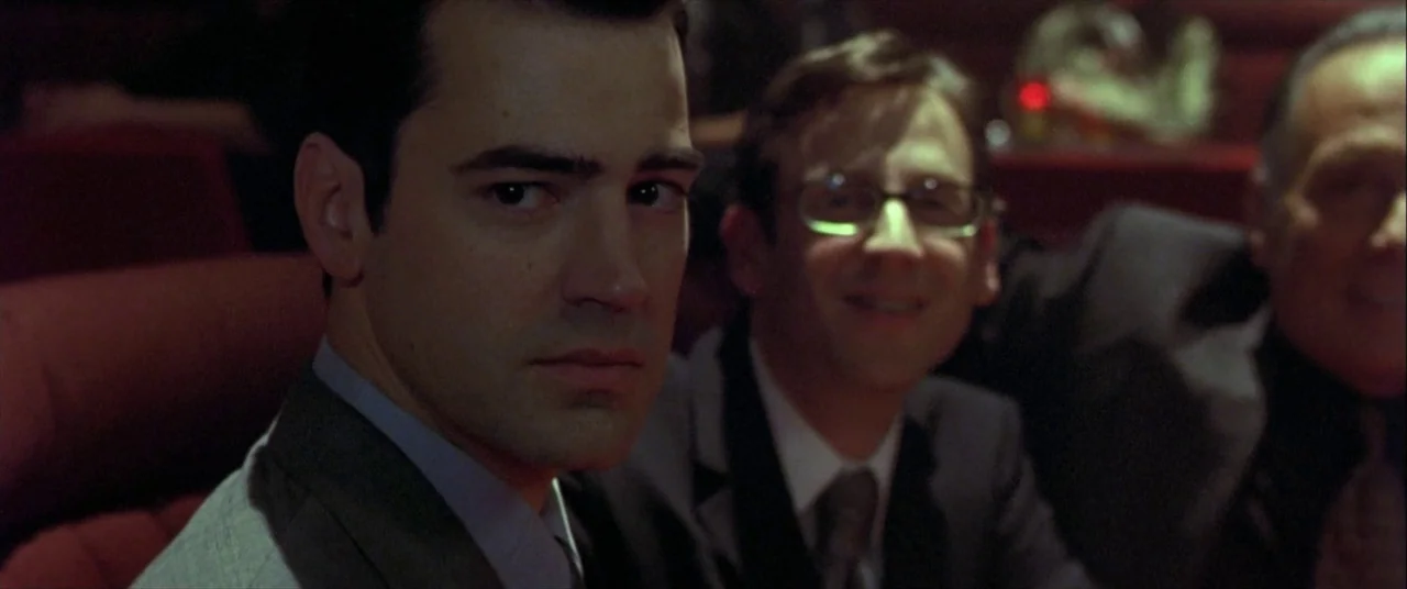The Cooler (2003) movie still: medium — Three men are seated in red velvet seats inside a dark public performance venue. The foreground subject…