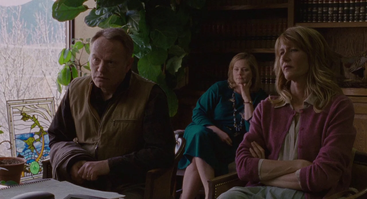 Certain Women (2016) movie still: medium — A dimly lit room contains wooden furniture, a large houseplant, and a small stained-glass piece. Three…