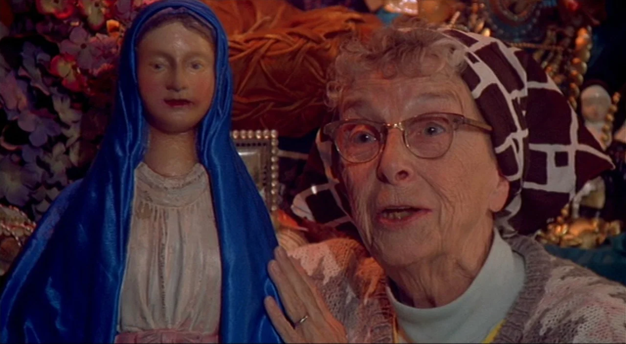 Pecker movie still: medium — An interior space filled with various trinkets and artificial flowers on display. An elderly woman in a headscarf…