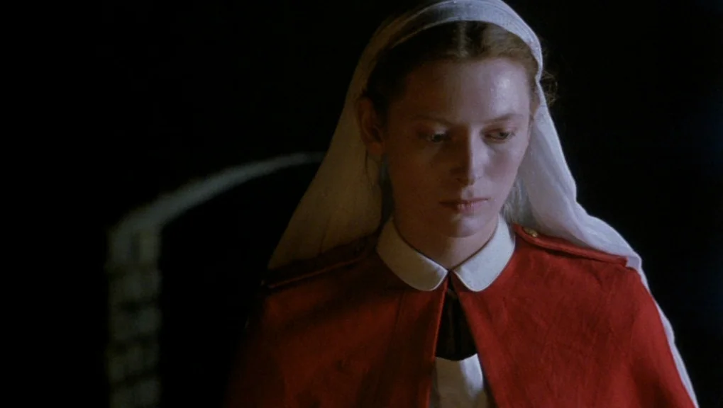 War Requiem (1989) movie still: closeup — A woman wears a 1940s-style nurse uniform with a red cape and headpiece. She stands against a dark,…
