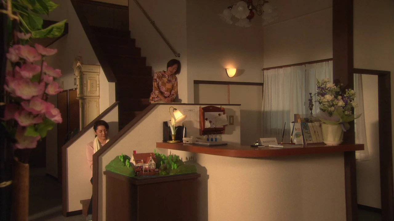 The Happiness of the Katakuris (2001) movie still: wide — A hotel lobby features a wooden reception desk and a staircase leading to upper floors.…