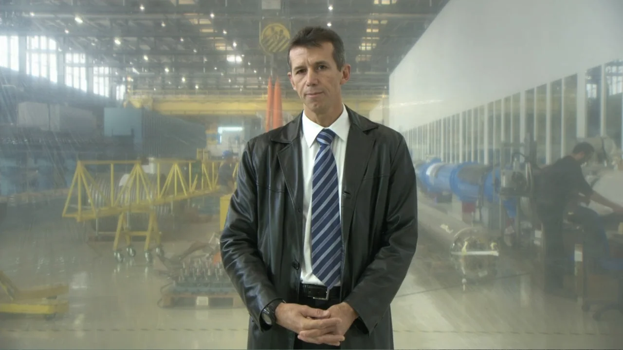 CERN (2013) movie still: medium — A spacious industrial manufacturing floor with high ceilings and metallic machinery in the background. A man in a…