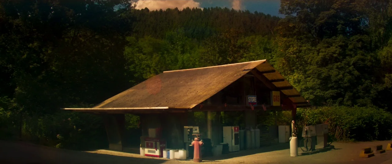 Mandy (2018) movie still: wide — An isolated wooden gas station structure sits nestled against a dense, forested hillside. The building features an…