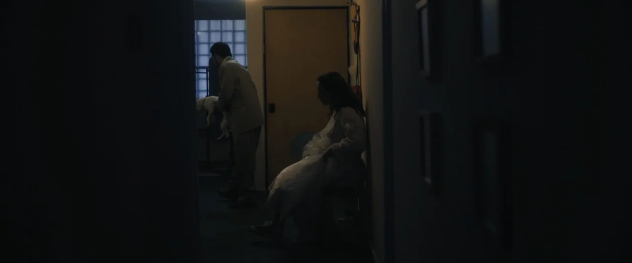 Sign Here (2017) movie still: medium — A dimly lit narrow passage with a door at the far end. A man holding a white dog stands near a seated woman…