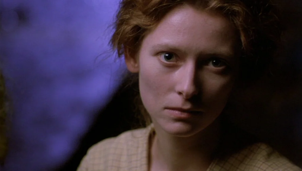 War Requiem (1989) movie still: closeup — A dark indoor space with a vibrant blue wall behind the subject. A woman stares toward the camera from a…