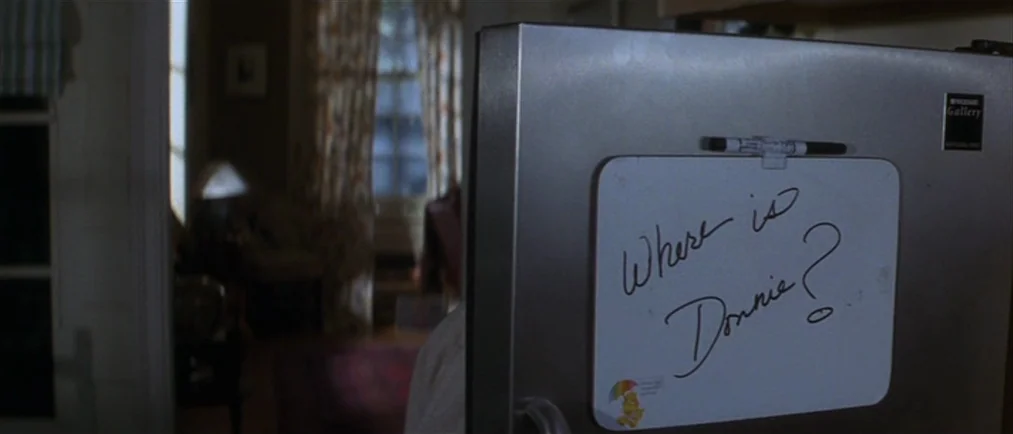 Donnie Darko movie still: closeup — A stainless steel refrigerator door features a dry-erase board with handwritten black ink. The blurred…
