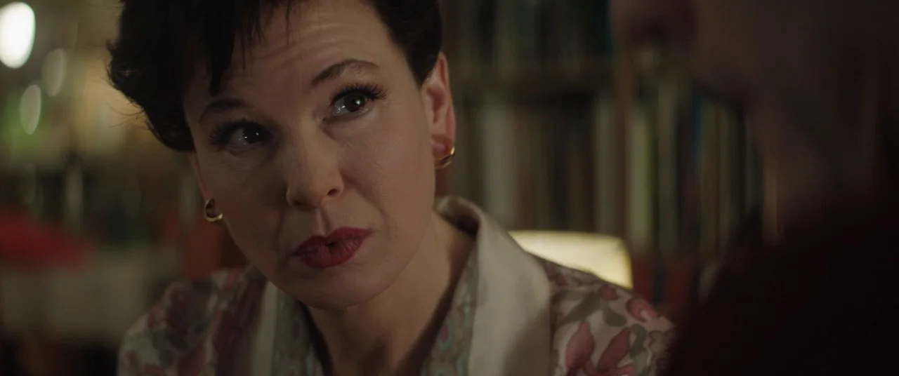Judy (2019) movie still: closeup — A woman with short dark hair looks toward a person in the foreground. Bookshelves and a soft glowing lamp are…
