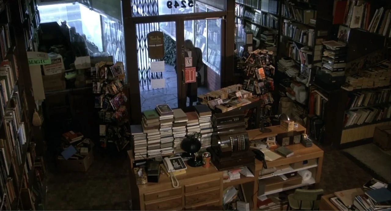 I, Madman (1989) movie still: wide — A cluttered bookstore features stacks of books, shelving units, and a wooden checkout counter. A lone man walks…