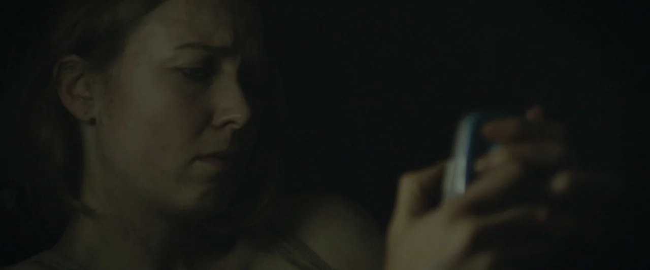 Here Alone movie still: closeup — A dimly lit room provides no environmental context beyond the darkness. A young woman holds a glowing smartphone…